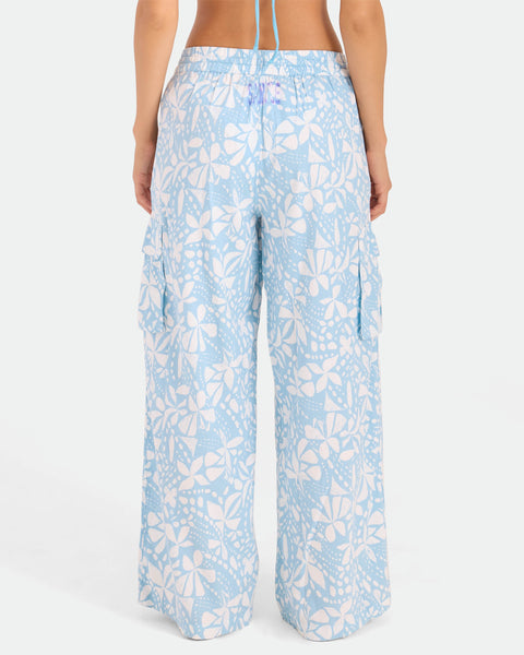 A person is seen from the back wearing Volcom Women's Wide Leg Pull On Pants in high-waisted light blue with a white floral and leaf print, soft cotton-viscose blend, side cargo pockets, elasticated waistband, and relaxed fit.