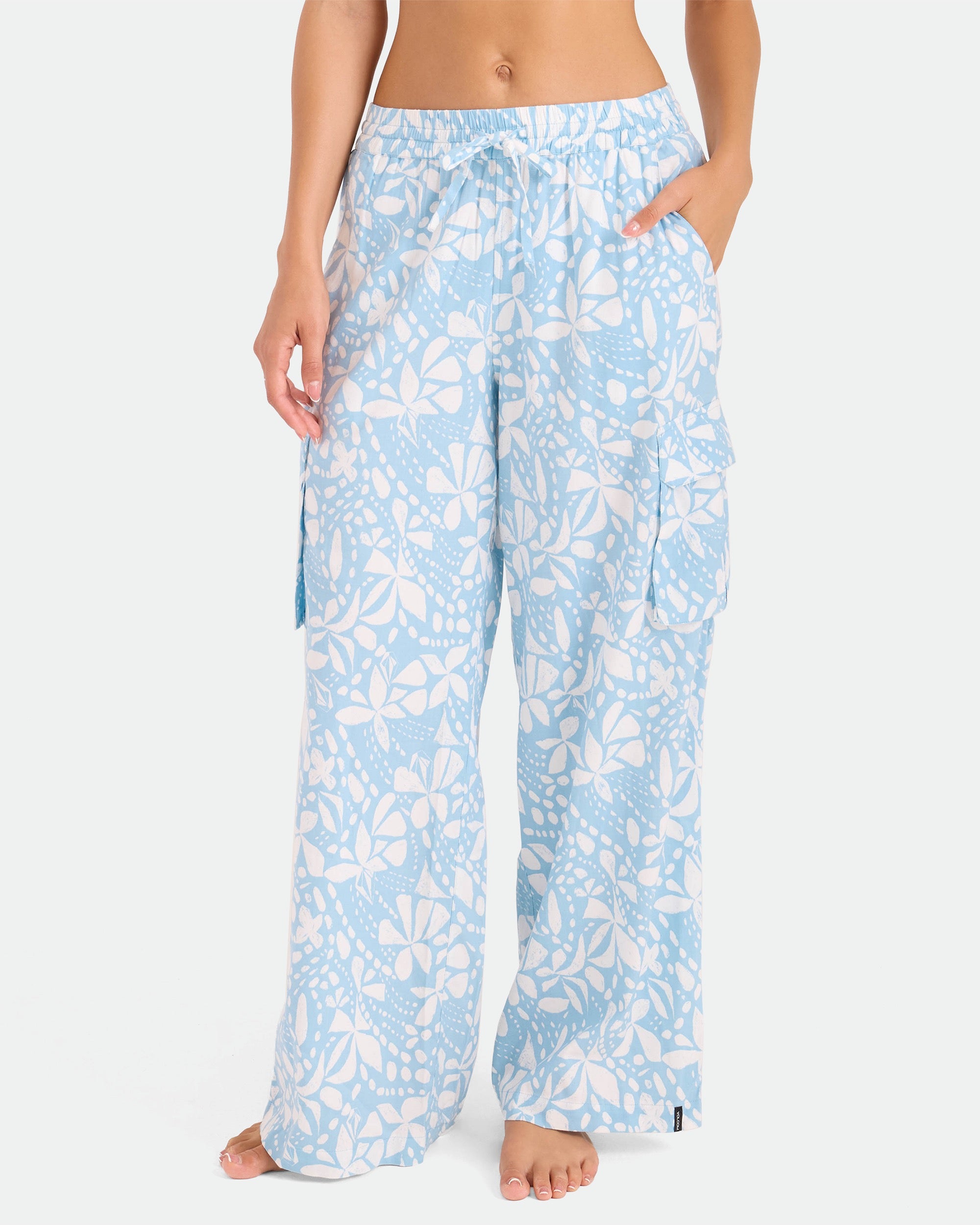 A woman stands barefoot against a plain white background, wearing Volcom Women's Wide Leg Pull On Pants in a light blue and white floral pattern with an elastic waistband and large side pockets.