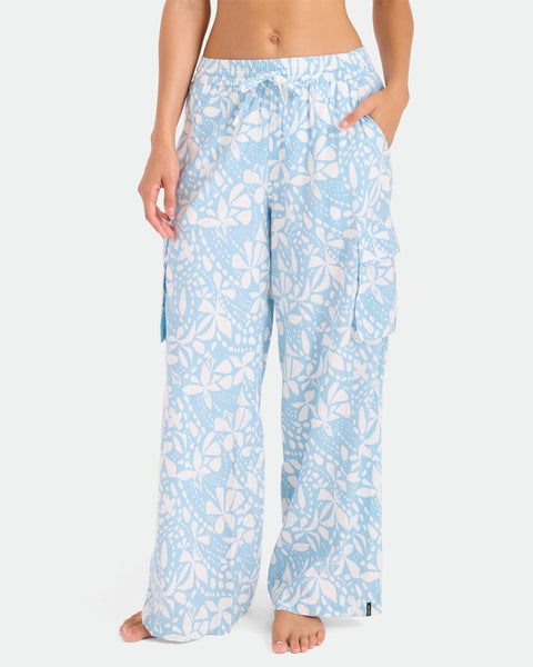A woman stands barefoot against a plain white background, wearing Volcom Women's Wide Leg Pull On Pants in a light blue and white floral pattern with an elastic waistband and large side pockets.
