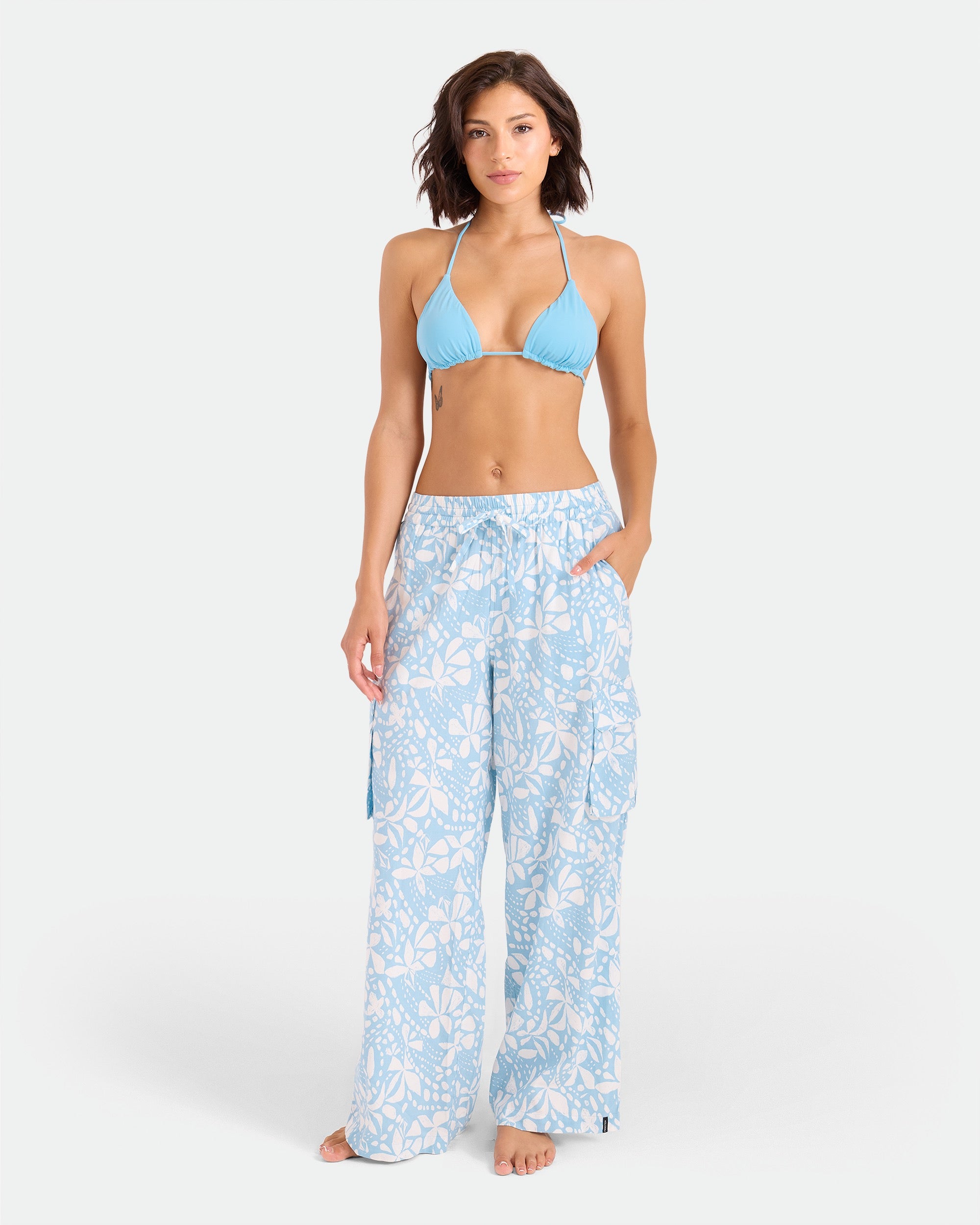 A woman stands barefoot against a white background, wearing a light blue bikini top and Volcom Women's Wide Leg Pull On Pants with a white floral pattern and elasticated waistband, her hands in her pockets.
