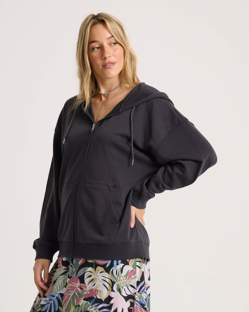 A young woman with long blonde hair poses against a plain background, wearing the Volcom Women’s Haleiwa Zip Hoodie in vintage black. The hoodie features soft fleece fabric, a hood, and practical pockets, and she pairs it with vibrant tropical-patterned pants while gazing slightly to the side.
