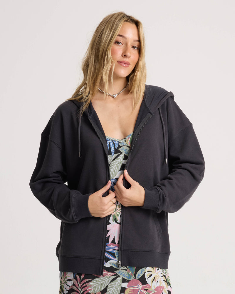 A woman wearing the Volcom Women’s Haleiwa Zip Hoodie in vintage black, celebrated for its cozy fleece feel, layers it over a vibrant, patterned dress as she stands against a plain background, gazing at the camera with a relaxed expression.