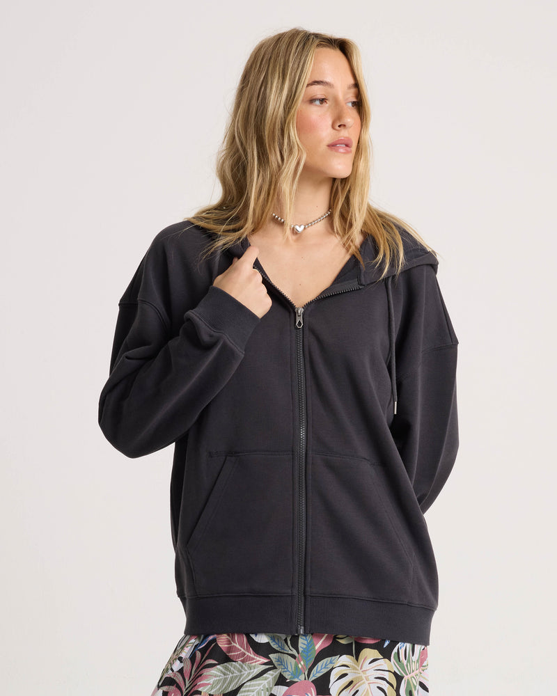 A woman with long blonde hair wears the Volcom Women’s Haleiwa Zip Hoodie, admired for its cozy fleece feel, effortlessly paired with a floral skirt. She stands against a plain white background, looking to the side.