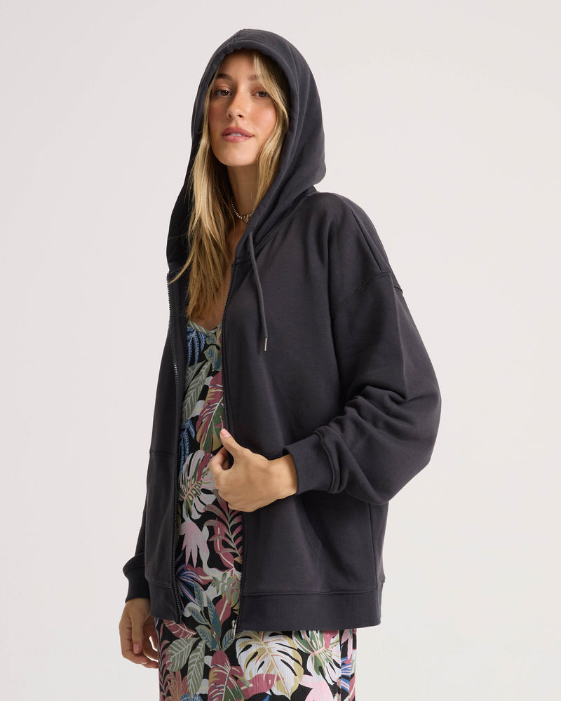 A person wears the Volcom Women’s Haleiwa Zip Hoodie in Vintage Black, layered over a vibrant tropical-print dress, standing against a plain light background and looking at the camera, showcasing its effortless fleece comfort.