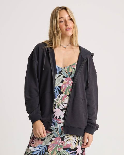 Clad in the Volcom Women’s Haleiwa Zip Hoodie in Vintage Black, renowned for its cozy fleece comfort, a woman stands against a plain light backdrop over a vibrant tropical print dress, gazing slightly upward with a relaxed expression.