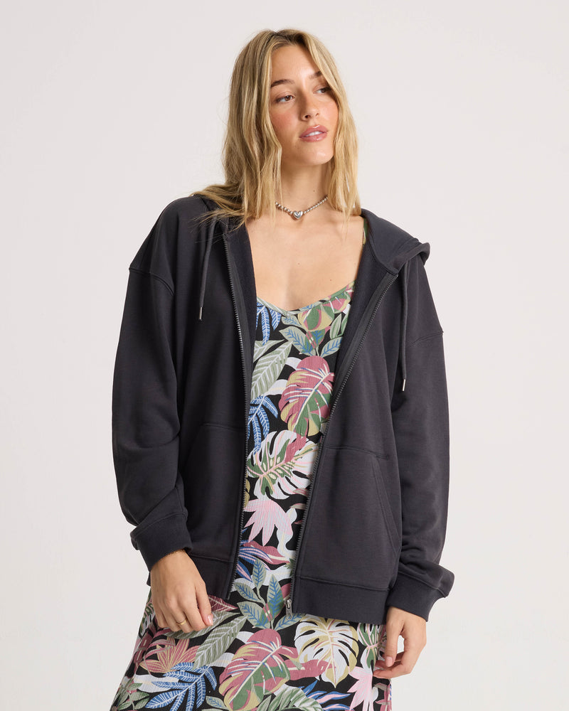 Clad in the Volcom Women’s Haleiwa Zip Hoodie in Vintage Black, renowned for its cozy fleece comfort, a woman stands against a plain light backdrop over a vibrant tropical print dress, gazing slightly upward with a relaxed expression.