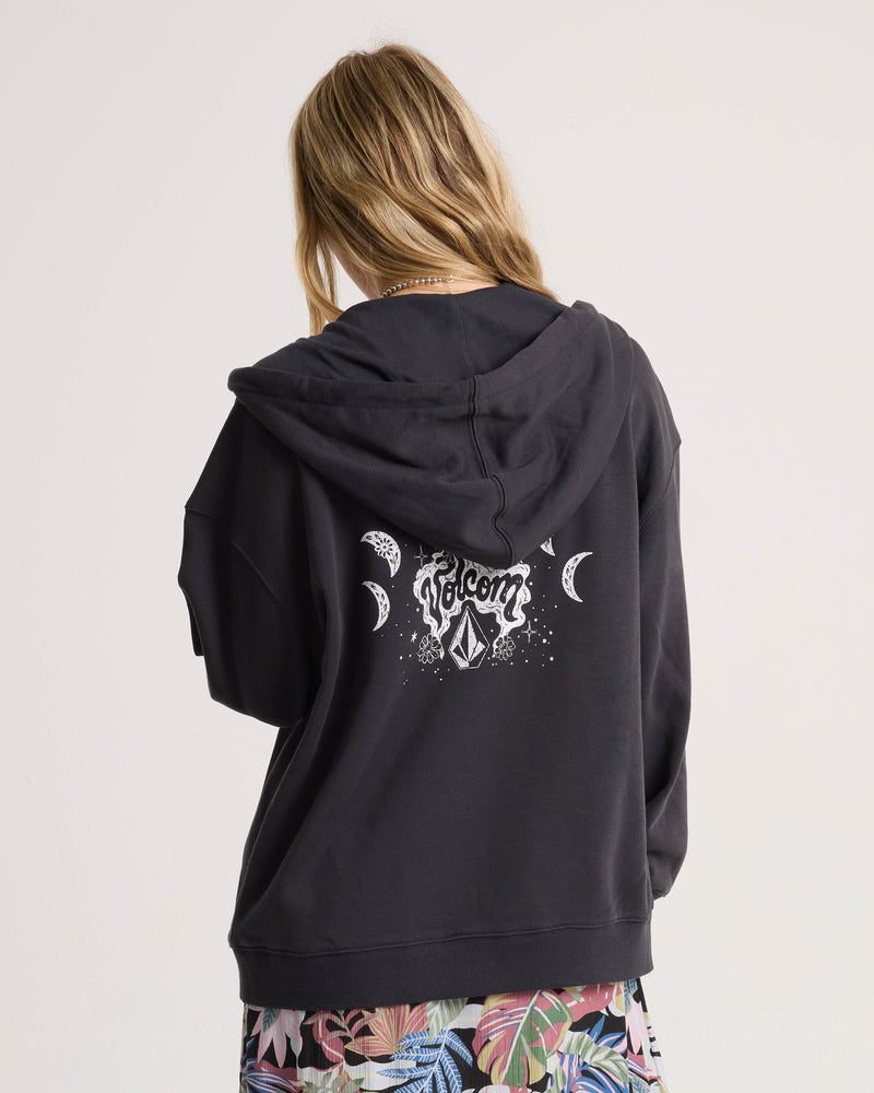 A woman with long hair faces away, wearing the Volcom Women’s Haleiwa Zip Hoodie in vintage black, featuring white celestial designs and “Volcom” on the back. She pairs it with vibrant, leafy print shorts for a cozy fleece look that stands out in bold style.