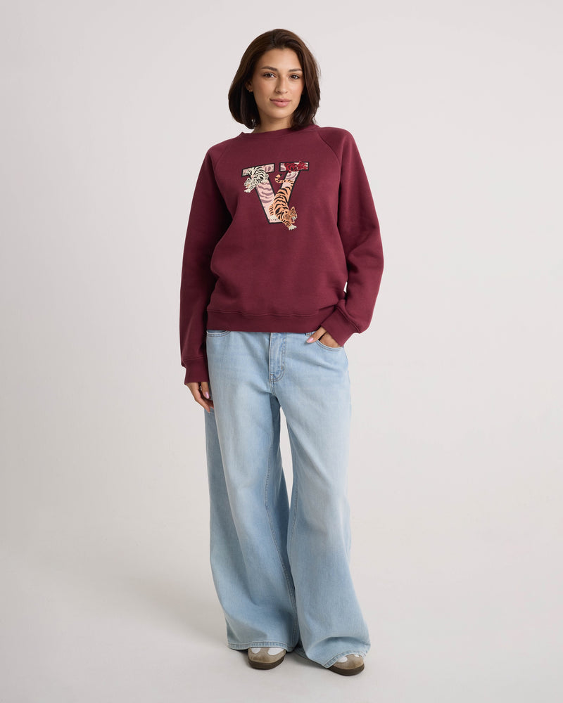 A woman wears the Volcom Women's Stone Magic BF maroon soft fleece pullover hoodie, which features a decorative letter V on the front. She pairs it with light blue wide-leg jeans and sneakers, standing against a plain light background. The relaxed fit of the hoodie brings effortless comfort to her casual look.