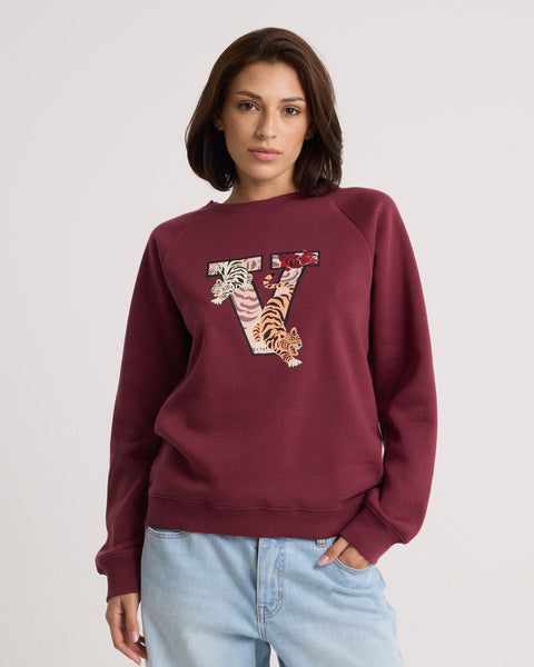A woman wears the Volcom Women's Stone Magic BF maroon pullover hoodie, featuring a decorative V and tiger design on the front and crafted from soft fleece for comfort, paired with light blue jeans and standing against a plain light background.