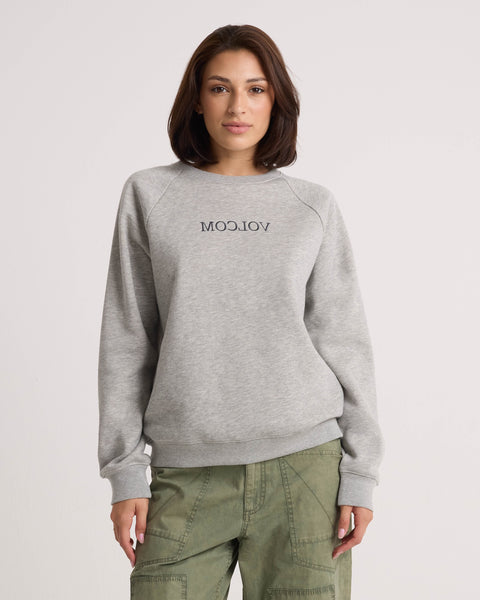 A woman stands against a plain light background with her hands by her sides, wearing the Volcom Women's Stone Magic BF pullover in heather gray and olive green pants. The relaxed fit of this sweatshirt adds to the laid-back vibe of her look.