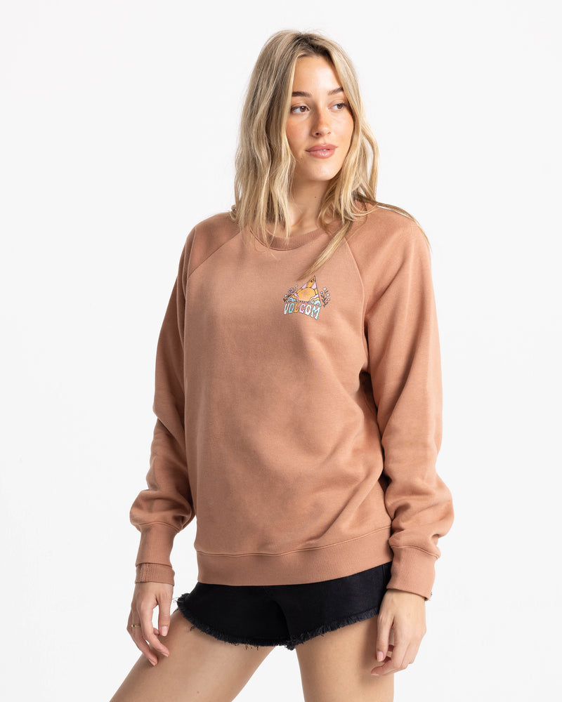 A woman with long blonde hair wears the Volcom Women’s Stone Magic Bf crew neck sweatshirt in Mocha Mousse, featuring a small embroidered design, paired with black shorts. She stands against a plain white background, looking to her right with a slight smile.