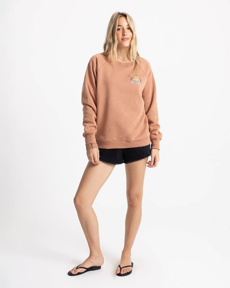 A woman wearing the Volcom Women’s Stone Magic Bf soft fleece crew neck sweatshirt in Mocha Mousse, featuring a small graphic on the chest, pairs it with black shorts and black sandals as she stands against a plain white background, smiling slightly.
