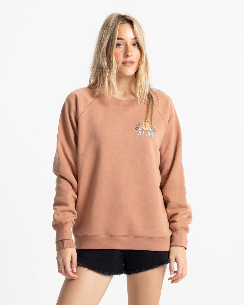 A woman with long blonde hair wears the Volcom Women’s Stone Magic Bf crew neck sweatshirt in Mocha Mousse, crafted from soft fleece, along with black shorts, standing against a plain white background.