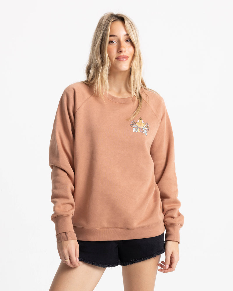 A woman with long blonde hair wears the Volcom Women’s Stone Magic Bf crew neck sweatshirt in tan, made from soft fleece, paired with black shorts as she stands against a plain white background.