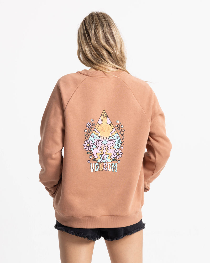 A person with long, wavy blonde hair is wearing the Women’s Stone Magic Bf crew neck sweatshirt by Volcom in a peach color, crafted from soft fleece with a colorful graphic and flower design on the back. The outfit is paired with black shorts and photographed against a white background.