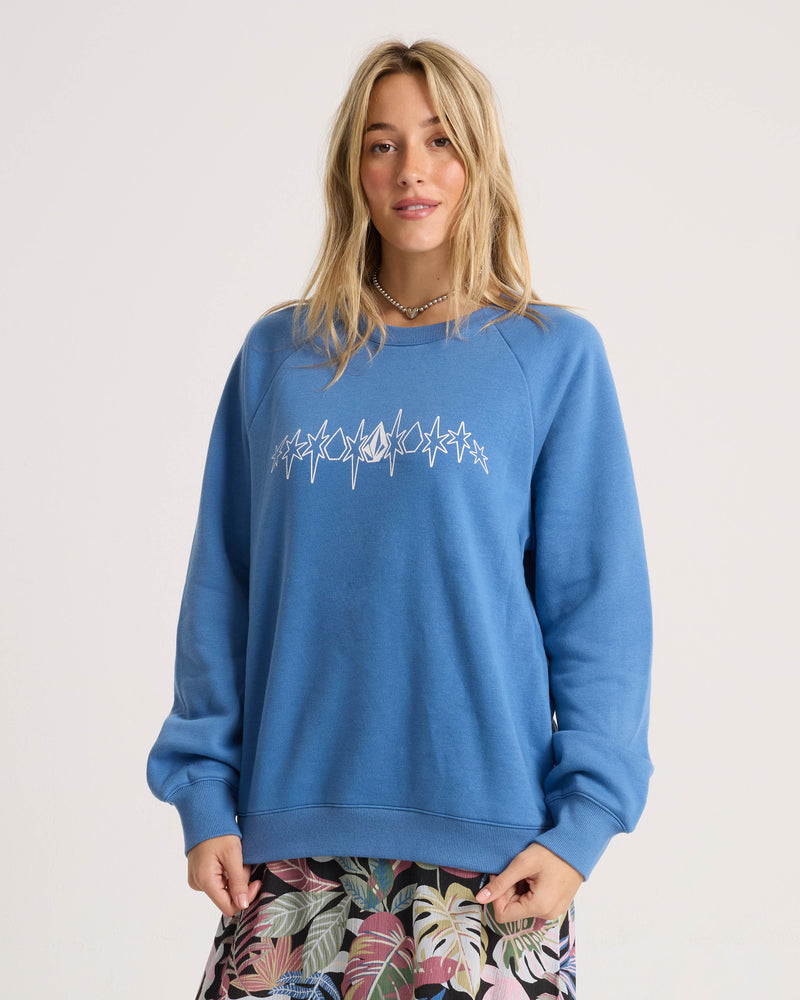 A woman with long blonde hair stands against a plain background, wearing the Volcom Women’s Stone Magic Bf sweatshirt in a relaxed fit blue style featuring a white abstract design. She pairs it with a patterned skirt adorned with leaf motifs.