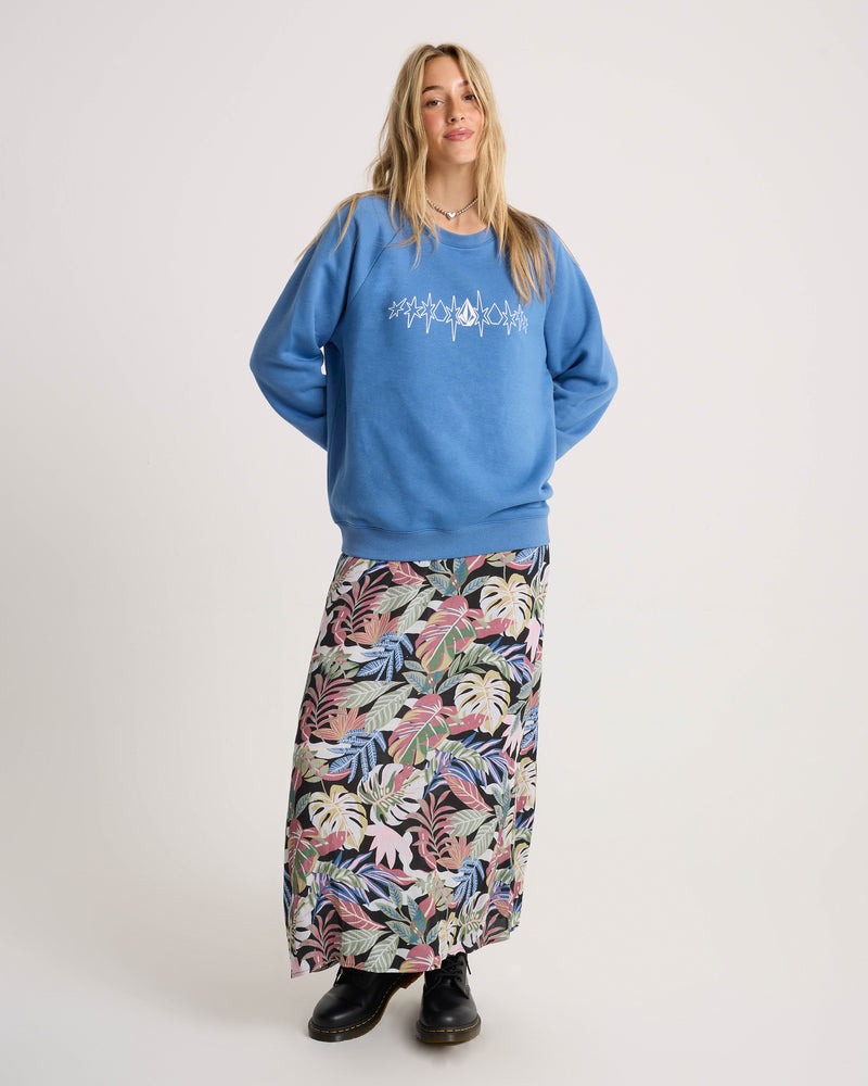 A woman with long blonde hair stands against a plain background, wearing the Volcom Women’s Stone Magic Bf sweatshirt in blue with a relaxed fit and white graphic on the front. She pairs it with a long, colorful leaf-patterned skirt and black shoes.