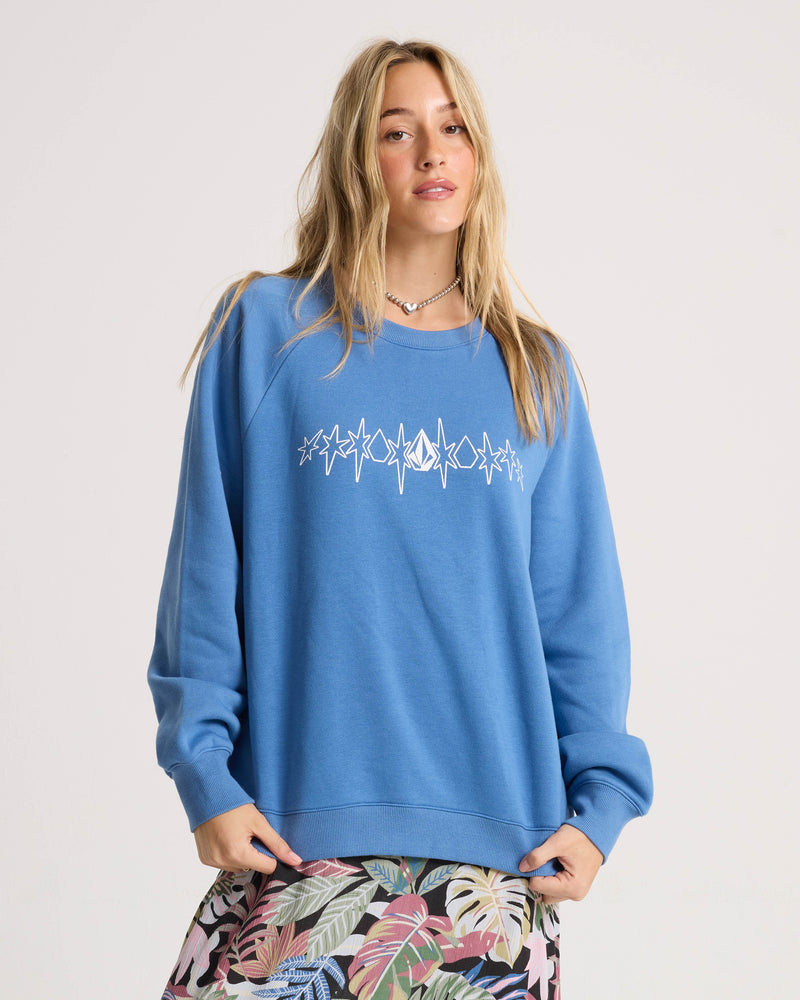 A woman with long blonde hair wears the Volcom Women’s Stone Magic Bf relaxed fit blue sweatshirt, which features a white geometric design, paired with a colorful skirt with leaf patterns, standing against a plain light background.