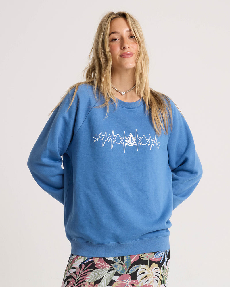 Woman with long blonde hair wears a relaxed fit, Riverside-hued Volcom Women’s Stone Magic Bf sweatshirt featuring white star designs, paired with a colorful leaf-patterned skirt, standing against a plain background.