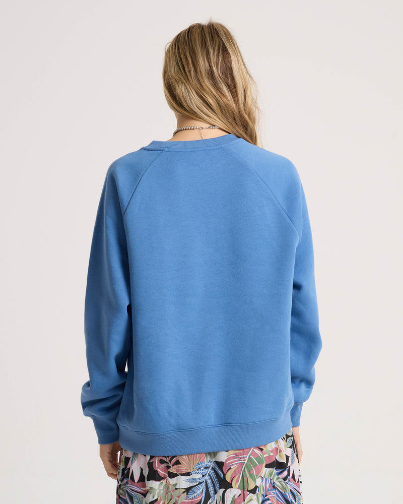 A person with long blonde hair, wearing the Volcom Women’s Stone Magic Bf sweatshirt in blue and a colorful floral skirt, stands facing away from the camera against a plain light background.