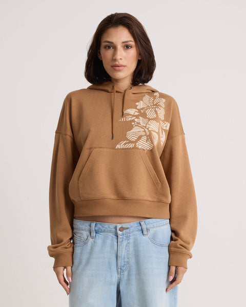 A woman with short brown hair stands facing forward in a Volcom Women's Stone Dream Hoodie—a brown relaxed-fit style featuring a white floral design on the shoulder. She pairs it with light blue jeans, set against a plain light-colored background.