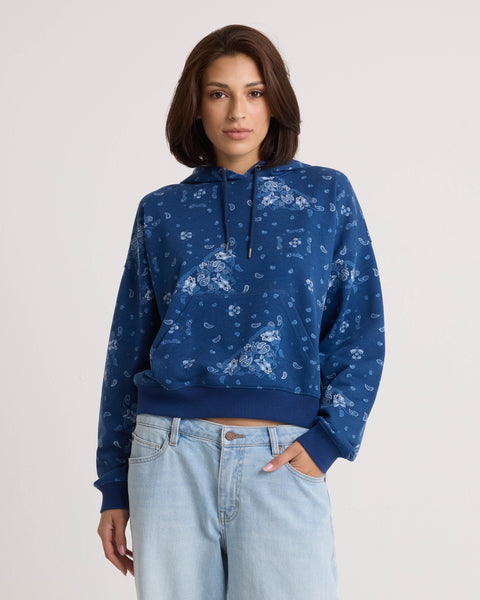 A woman wears the Volcom Women's Stone Dream Hoodie, featuring a blue relaxed fit with a light floral pattern, paired with light blue jeans. She stands against a plain light background, looking at the camera with a neutral expression.