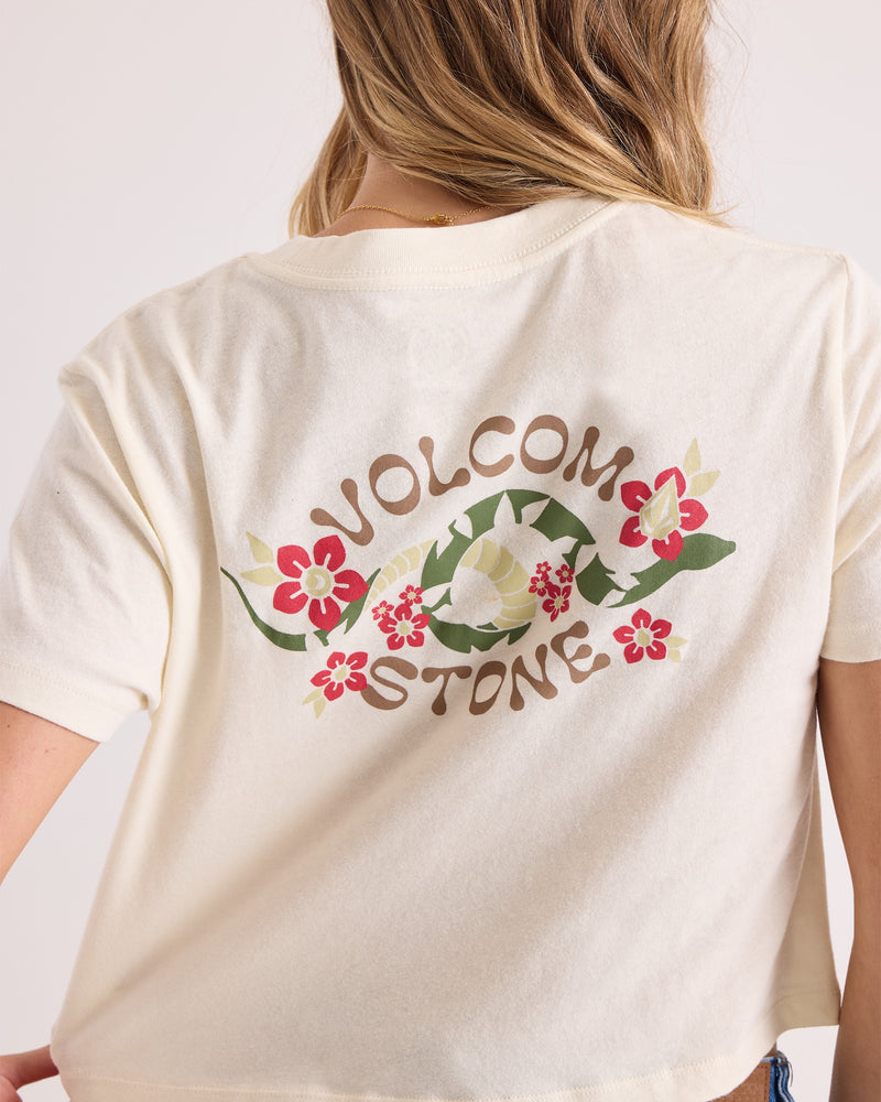 A woman wears the Volcom Women's Bite Club Pocket Dial Tee, a cream-colored short-sleeve knit top featuring "VOLCOM STONE" and floral graphics on the back. Only her back and partial arm are visible.
