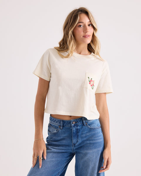 A woman wearing the Volcom Women's Bite Club Pocket Dial Tee—a cream-colored, 100% cotton cropped shirt featuring a floral design on the pocket—paired with blue jeans, stands against a plain light background.