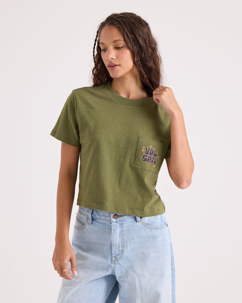 A woman wearing the Volcom Women's Super Melt Pocket Dial Tee in green—made from 100% cotton with a crew neck and pocket—and light blue jeans stands against a plain background, looking down and touching her hair.