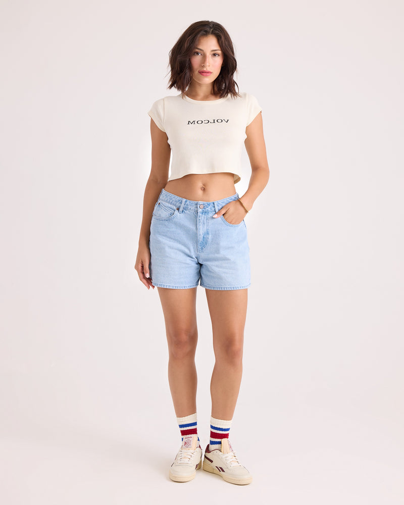 A woman stands against a plain background wearing the Volcom Women's Flip Side 1991 Mid Tee—a vintage-inspired, fitted white cropped shirt—paired with light blue denim shorts, white sneakers, and white socks featuring red and blue stripes.
