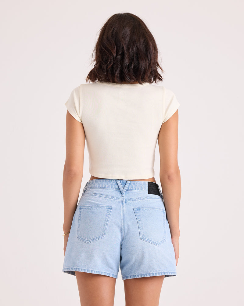 A woman with short dark hair is standing with her back to the camera, wearing the Volcom Women's Flip Side 1991 Mid Tee in vintage white and light blue denim shorts against a plain light background.