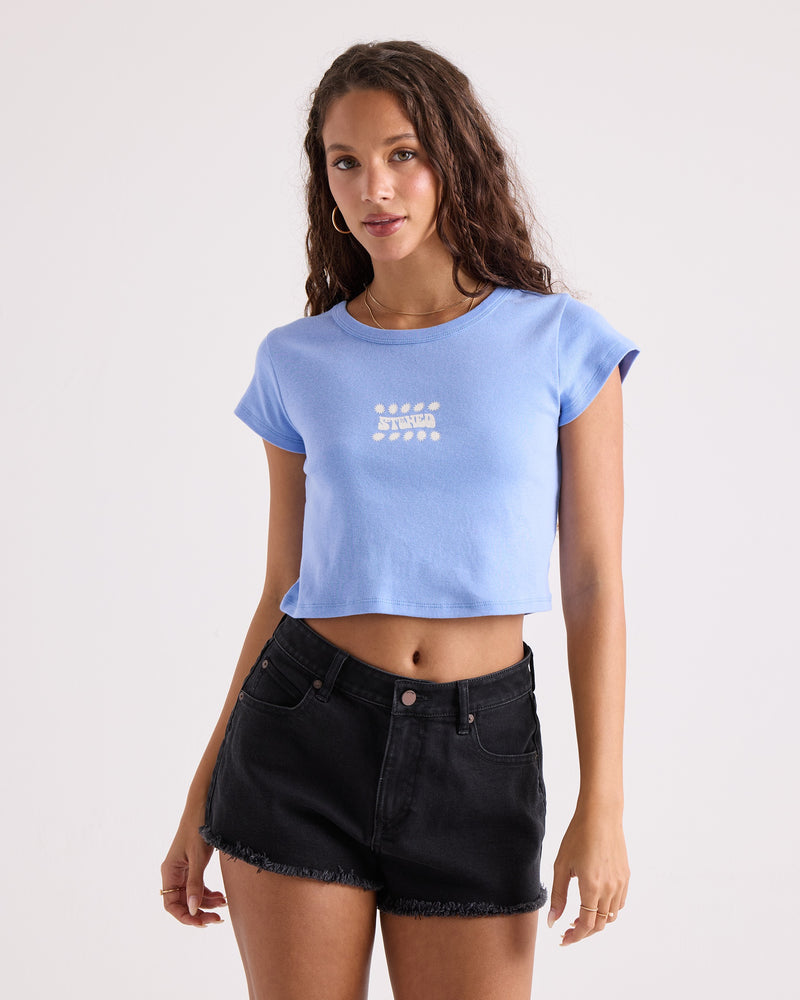A woman with long curly hair wears the Volcom Women's Detour 1991 Mid Tee—a blue, 100% cotton, cropped fitted t-shirt with white text—paired with black denim shorts, standing against a plain light background.