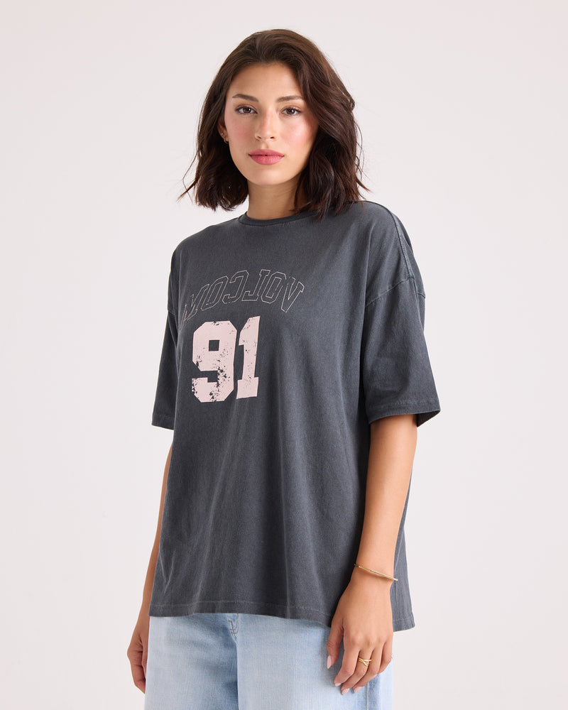 A woman with shoulder-length dark hair wears the Volcom Women's Line Up Base Tee, a loose-fitting black oversized t-shirt made from 100% cotton, featuring “MOODJOY 91” in white text. She pairs it with light blue jeans and stands against a plain light background.