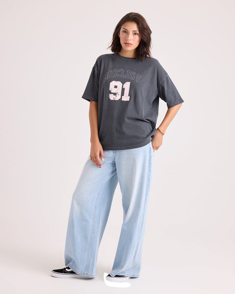 A woman wearing a Volcom Women's Line Up Base Tee in vintage black, crafted from 100% cotton, and light blue wide-leg jeans stands with one hand in her pocket against a plain background.