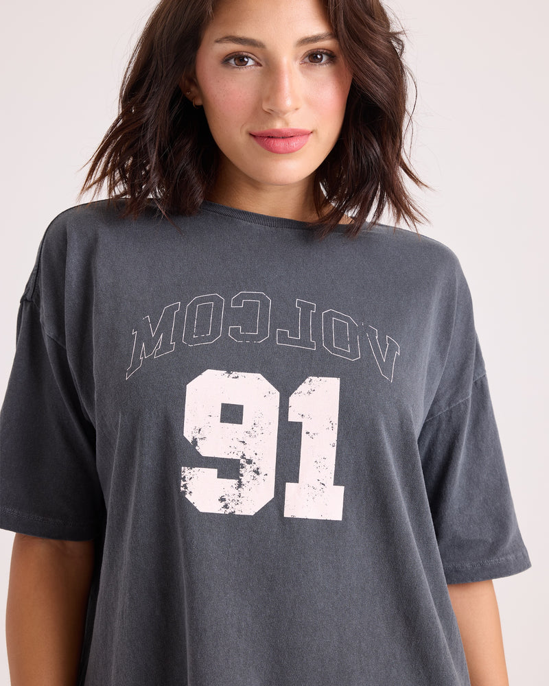 A woman with shoulder-length dark hair is wearing the Volcom Women's Line Up Base Tee, an oversized vintage black t-shirt featuring "VOLCOM 91" in white lettering. She stands against a plain background and smiles softly at the camera.