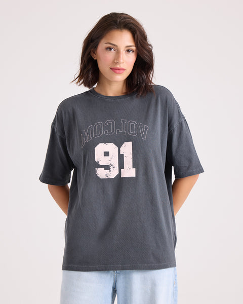 A woman stands against a plain background, hands behind her back and looking at the camera, wearing light blue jeans and the Volcom Women's Line Up Base Tee—a vintage black oversized t-shirt with "VOLCOM 91" in distressed white letters.