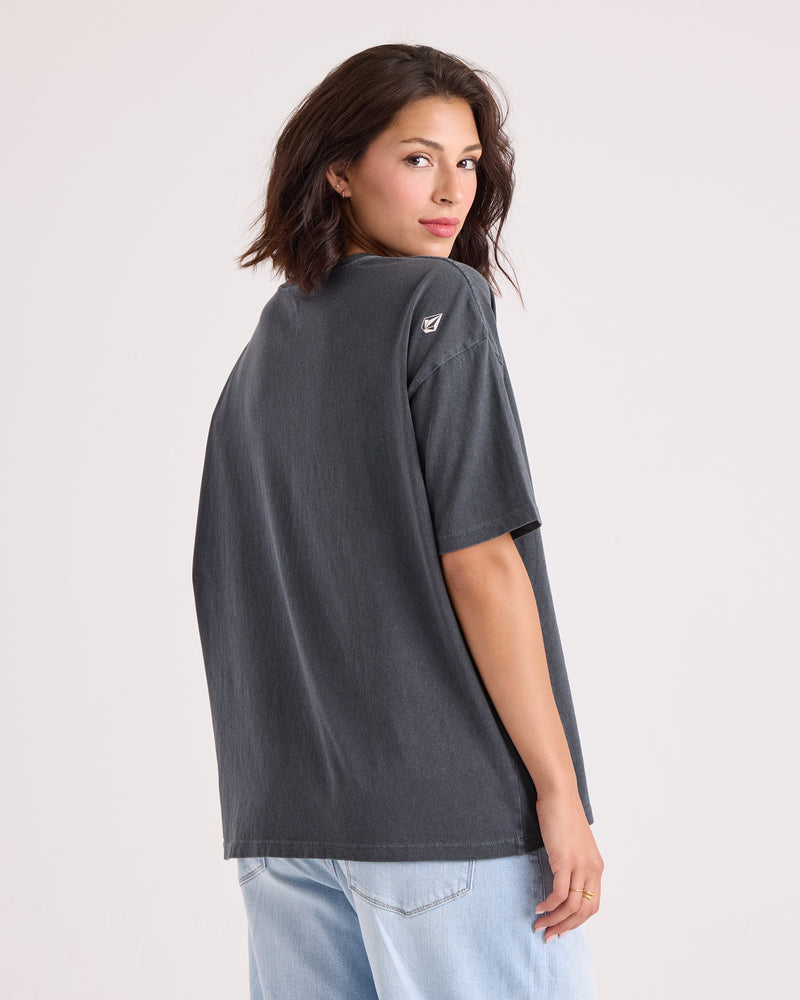 A woman with shoulder-length dark hair, wearing the Volcom Women's Line Up Base Tee—a vintage-inspired, oversized black t-shirt made from 100% cotton—paired with light blue jeans, stands turned slightly away from the camera and looks back over her shoulder against a plain background.