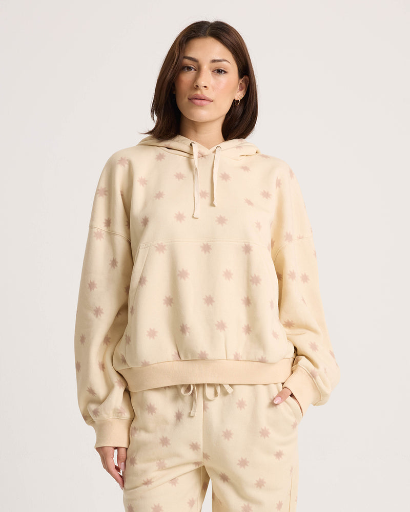 A woman wears the Volcom Women’s In A Cinch Sweatshirt and matching beige sweatpants with a light pink star print, standing facing forward with one hand in her pocket against a white background.