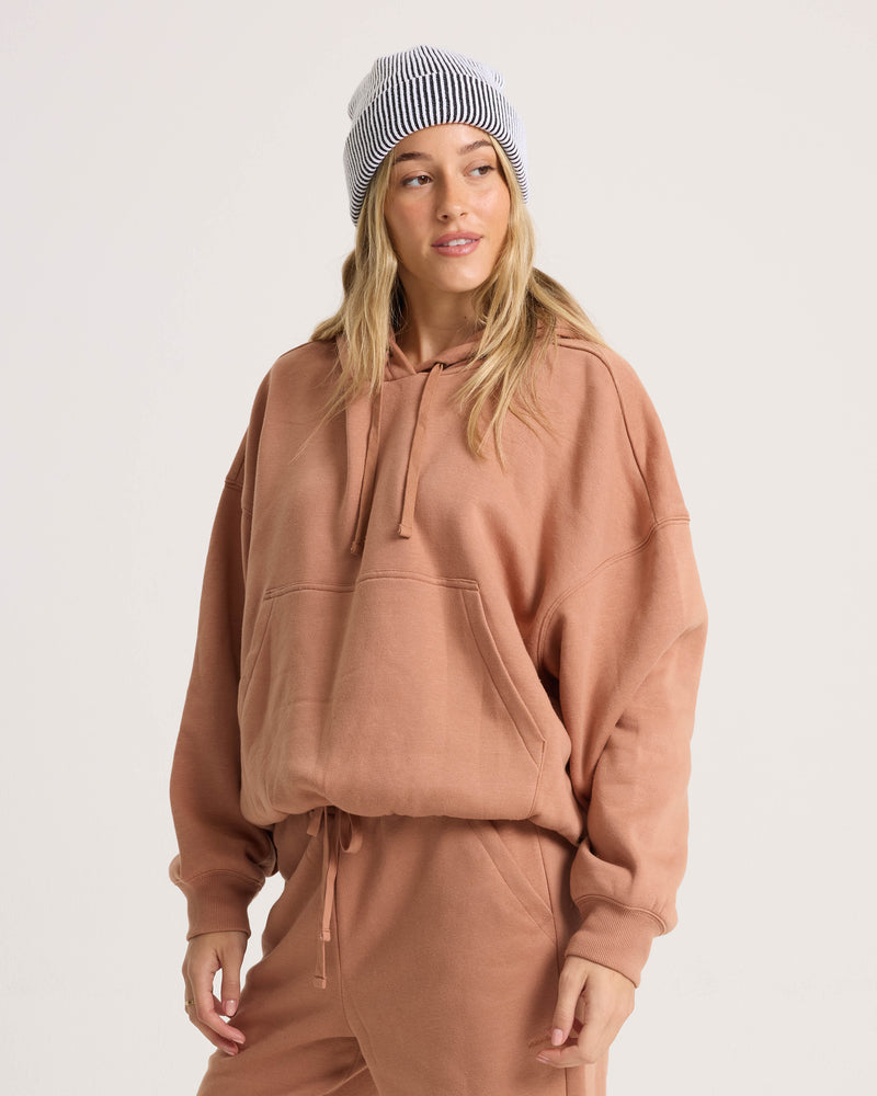 A woman in a Volcom Women’s In A Cinch Sweatshirt and matching sweatpants, paired with a white and black striped beanie, stands against a plain light background, gazing slightly to her left.