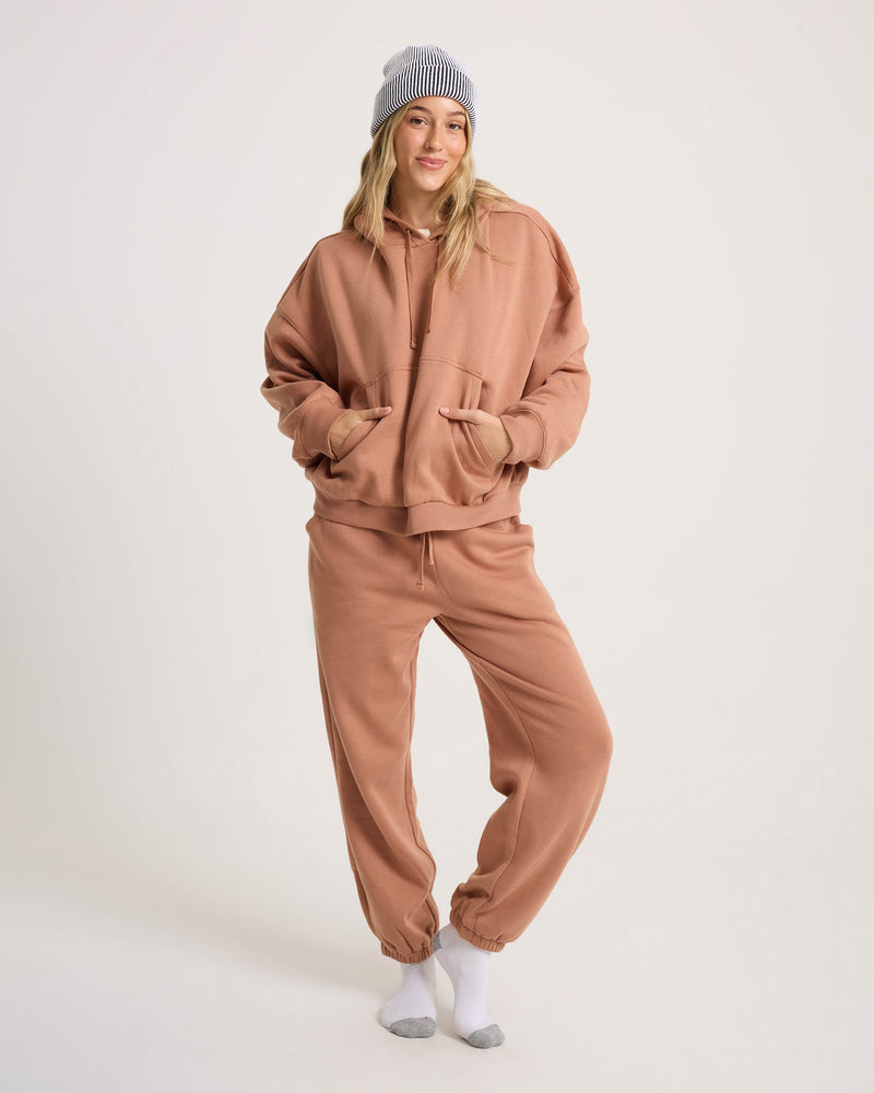 A person in a Volcom Women’s In A Cinch Sweatshirt and matching sweatpants stands on one leg, smiling with hands in pockets. They wear a striped beanie, long hair, and socks against a plain white background.