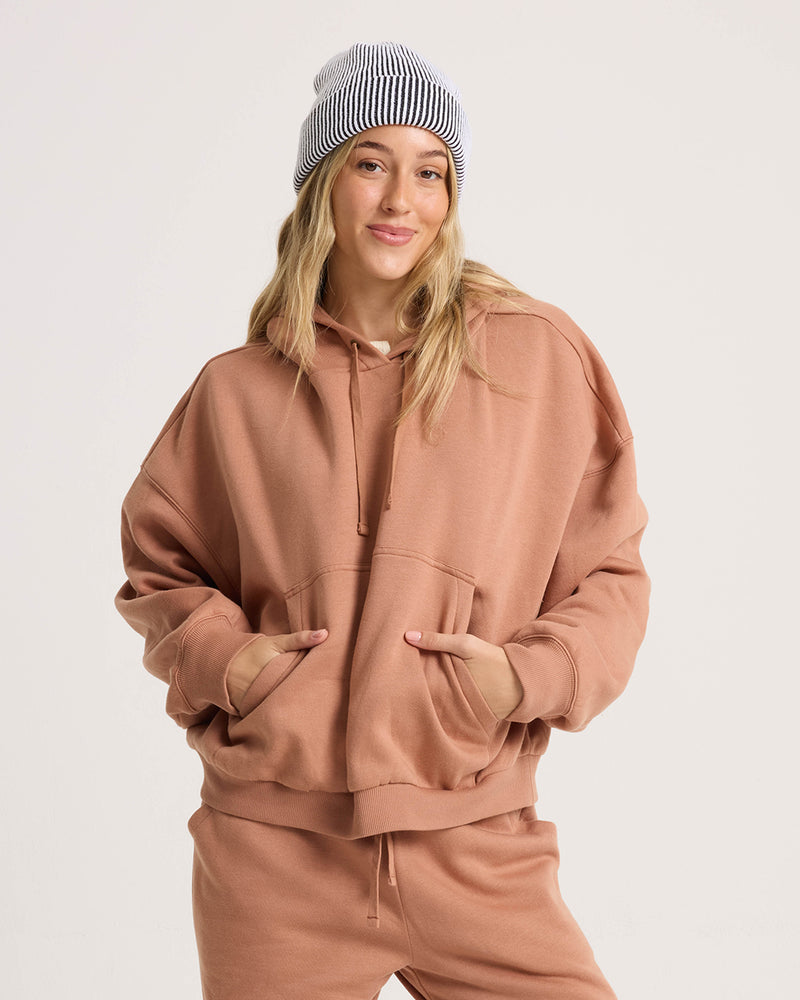 A person in a Volcom Women’s In A Cinch Sweatshirt and matching sweatpants stands with hands in the hoodie pocket, smiling slightly. They have long hair, a white and black striped beanie, and a light plain background.