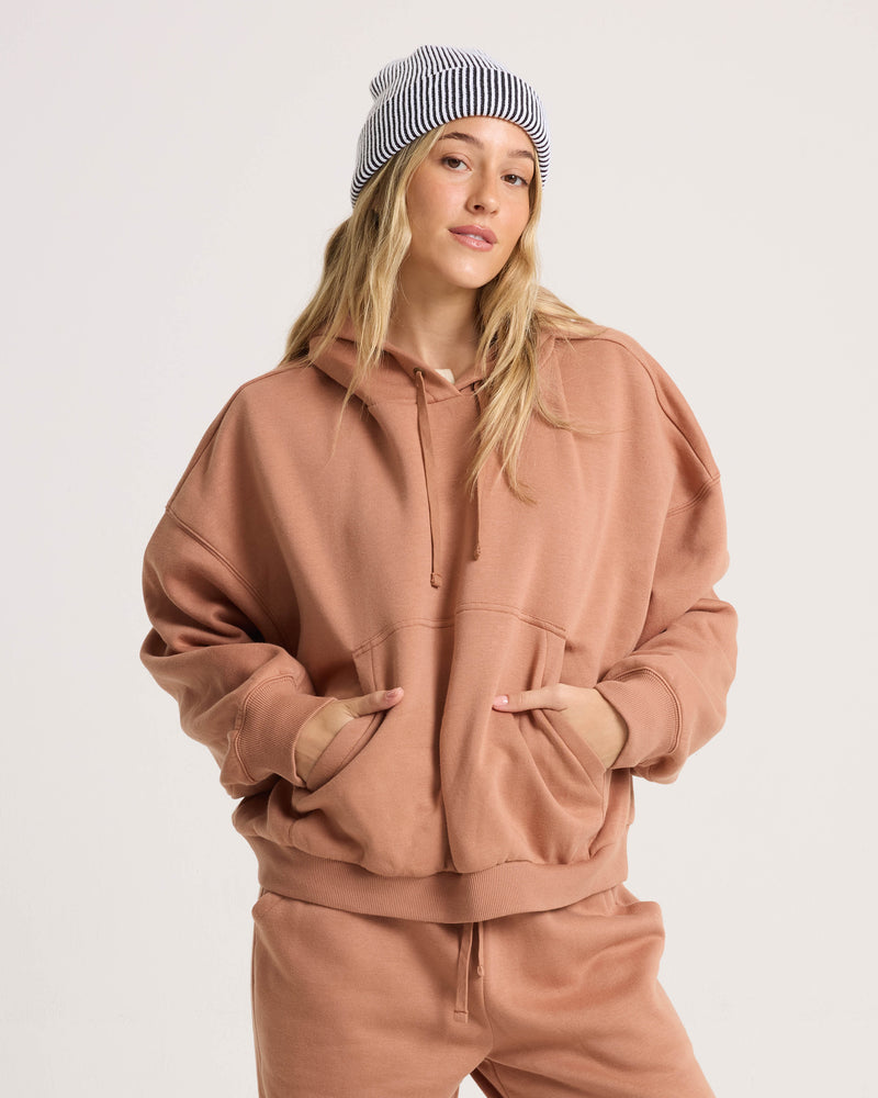 A woman in the Volcom Women’s In A Cinch Sweatshirt, a mocha mousse brown oversized pullover hoodie with matching sweatpants, poses confidently with long blonde hair and a white and black striped beanie.