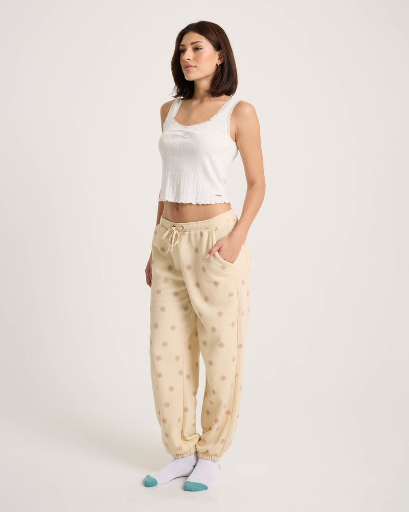 A woman wears a white tank top, Volcom Women’s In A Cinch Jogger Pant in beige with a subtle pattern, and white socks with blue toes, standing sideways with one hand in her pocket against a plain background.