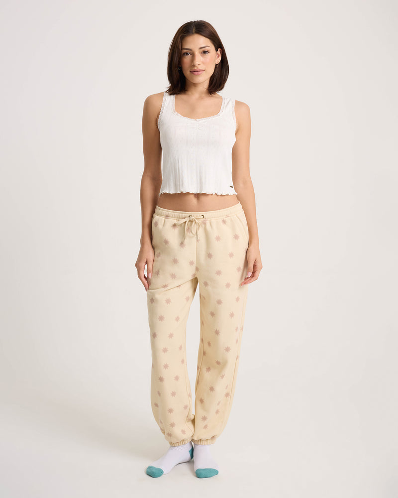 A woman stands facing forward, barefoot on a white background, wearing a white sleeveless crop top and Volcom Women’s In A Cinch Jogger Pant in beige with a small star pattern.