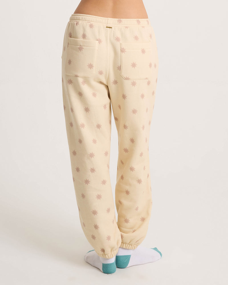 A person stands with feet crossed in Volcom Women’s In A Cinch Jogger Pant—loose fit, cream with a light pink star pattern, oversized with two back pockets, elastic cuffs, and paired with white and teal socks on a plain background.