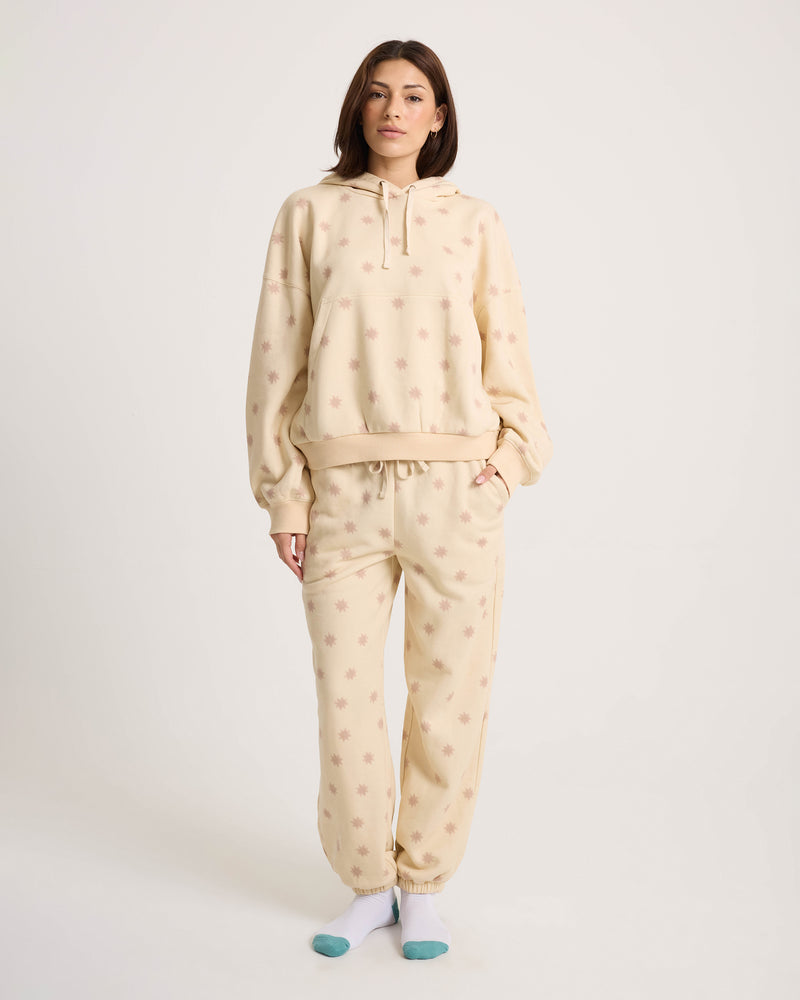 A woman stands facing forward, wearing the Volcom Women’s In A Cinch Jogger Pant in beige with a matching loose fit hoodie and mismatched socks against a plain white background.