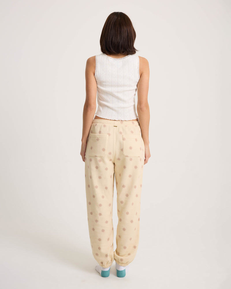 A person faces away from the camera, wearing a white sleeveless top and Volcom Women’s In A Cinch Jogger Pant in cream with a pink pattern against a plain white background.