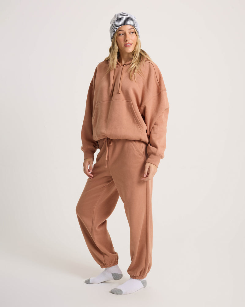 A person in a light brown oversized hoodie, Women’s In A Cinch Jogger Pant by Volcom, white socks, and a gray beanie stands against a plain background, showcasing everyday comfort.