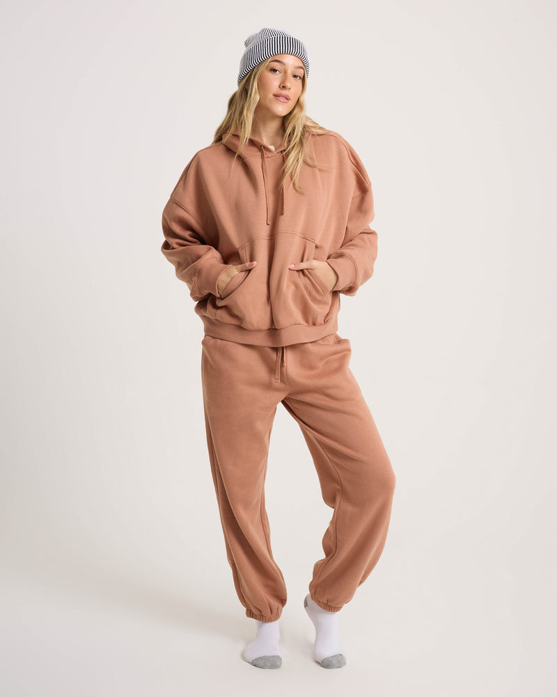 A person wears the Volcom Women’s In A Cinch Jogger Pant with a tan oversized hoodie, gray-and-white striped beanie, and white socks, standing against a white background with hands in hoodie pockets.