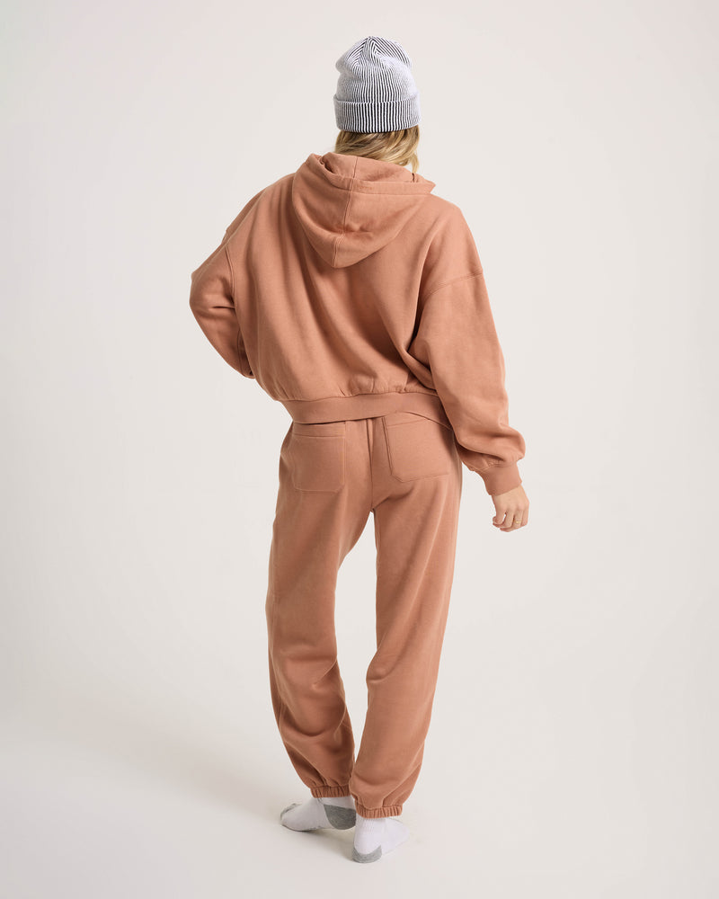 A person faces away, wearing an oversized light brown hoodie, Volcom Women’s In A Cinch Jogger Pant, a gray-and-white beanie, and white socks. The plain light background highlights comfy everyday style.